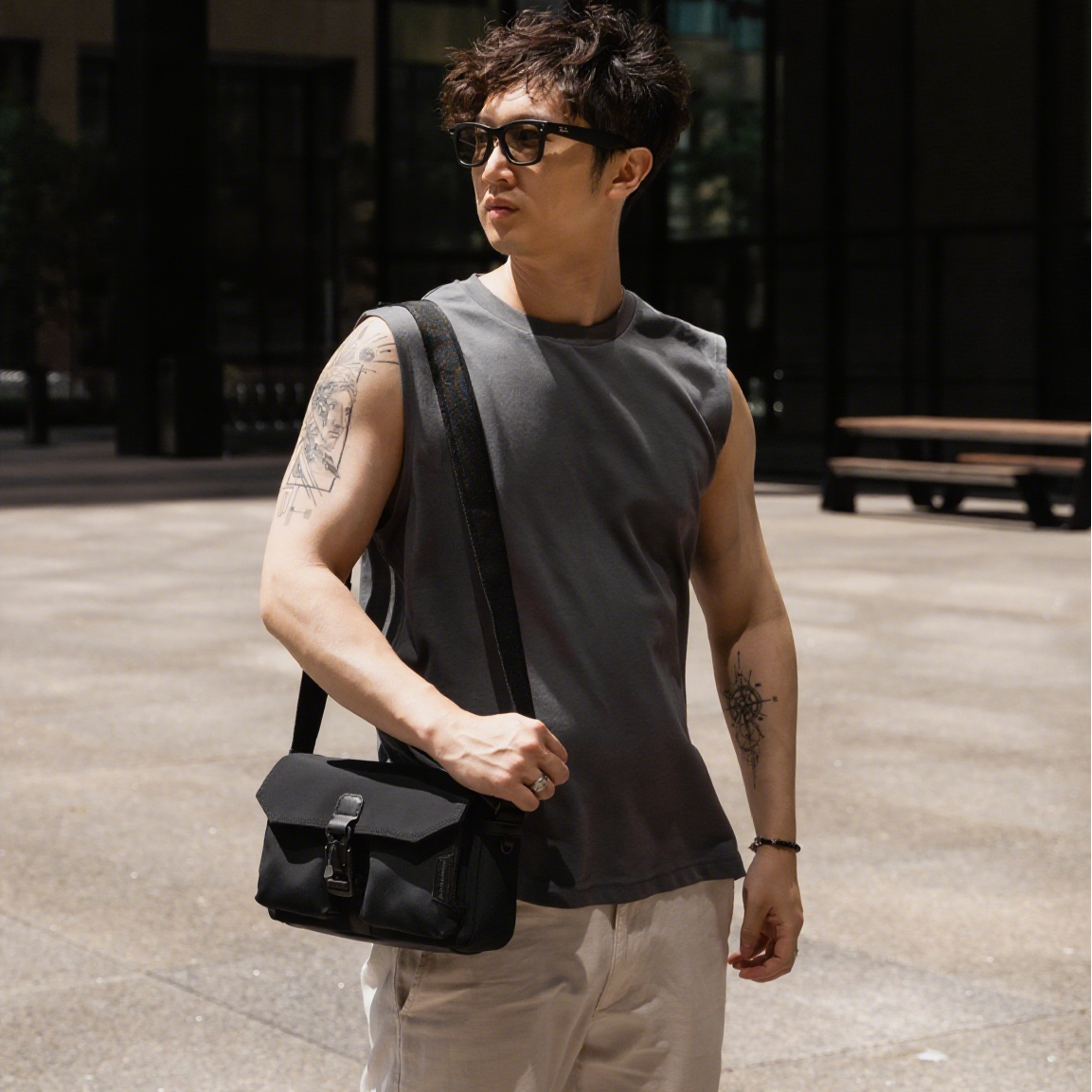 Man wearing sunglasses and a sleeveless shirt, holding a black bag in an urban setting.