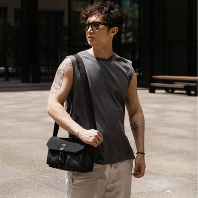 Man wearing sunglasses and a sleeveless shirt, holding a black bag in an urban setting.