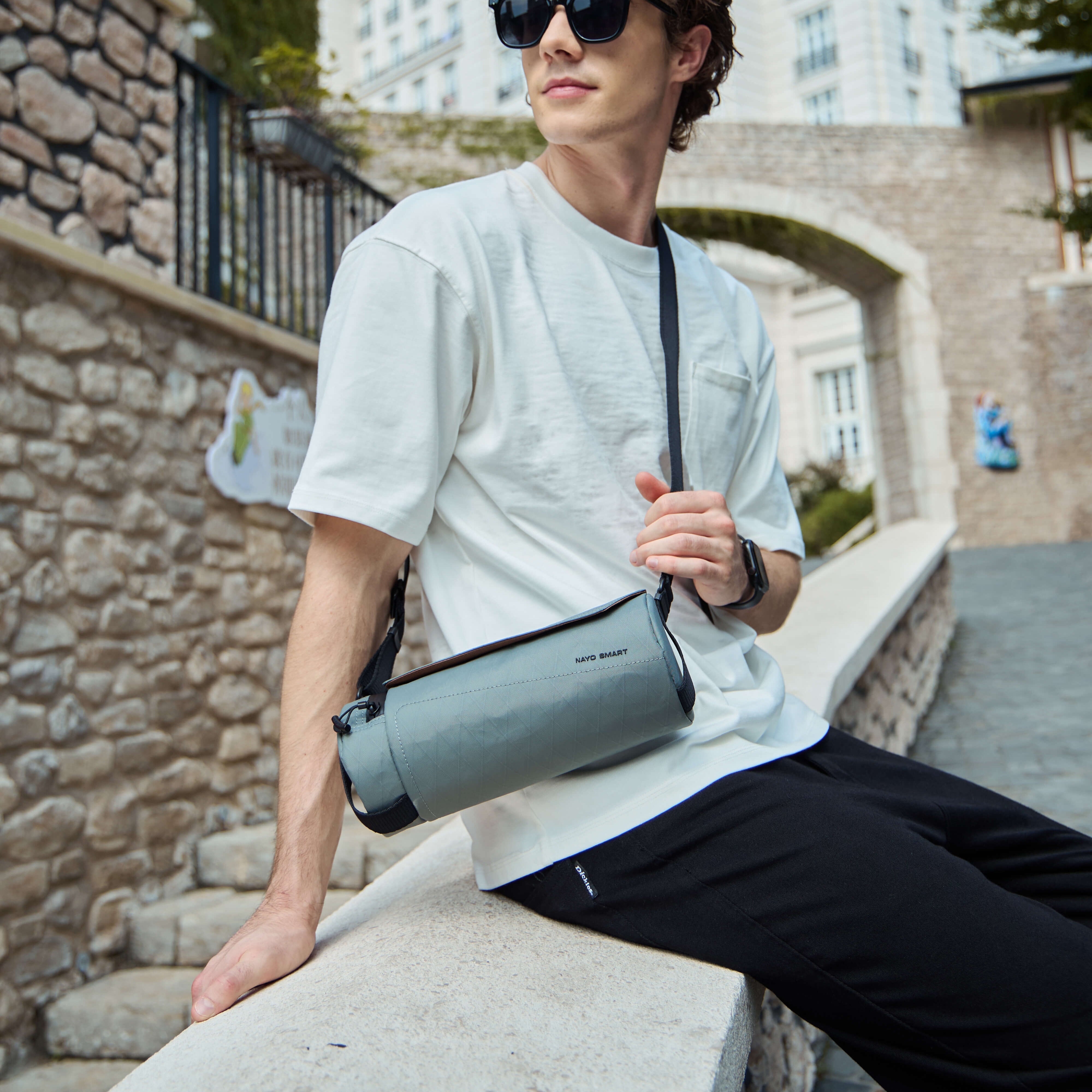 Outdoor European city lifestyle shot, man wearing white tee carrying grey X‑Pac Nayo Smart UL5 Cylinder Sling Bag, compact barrel‑shaped city crossbody travel bag