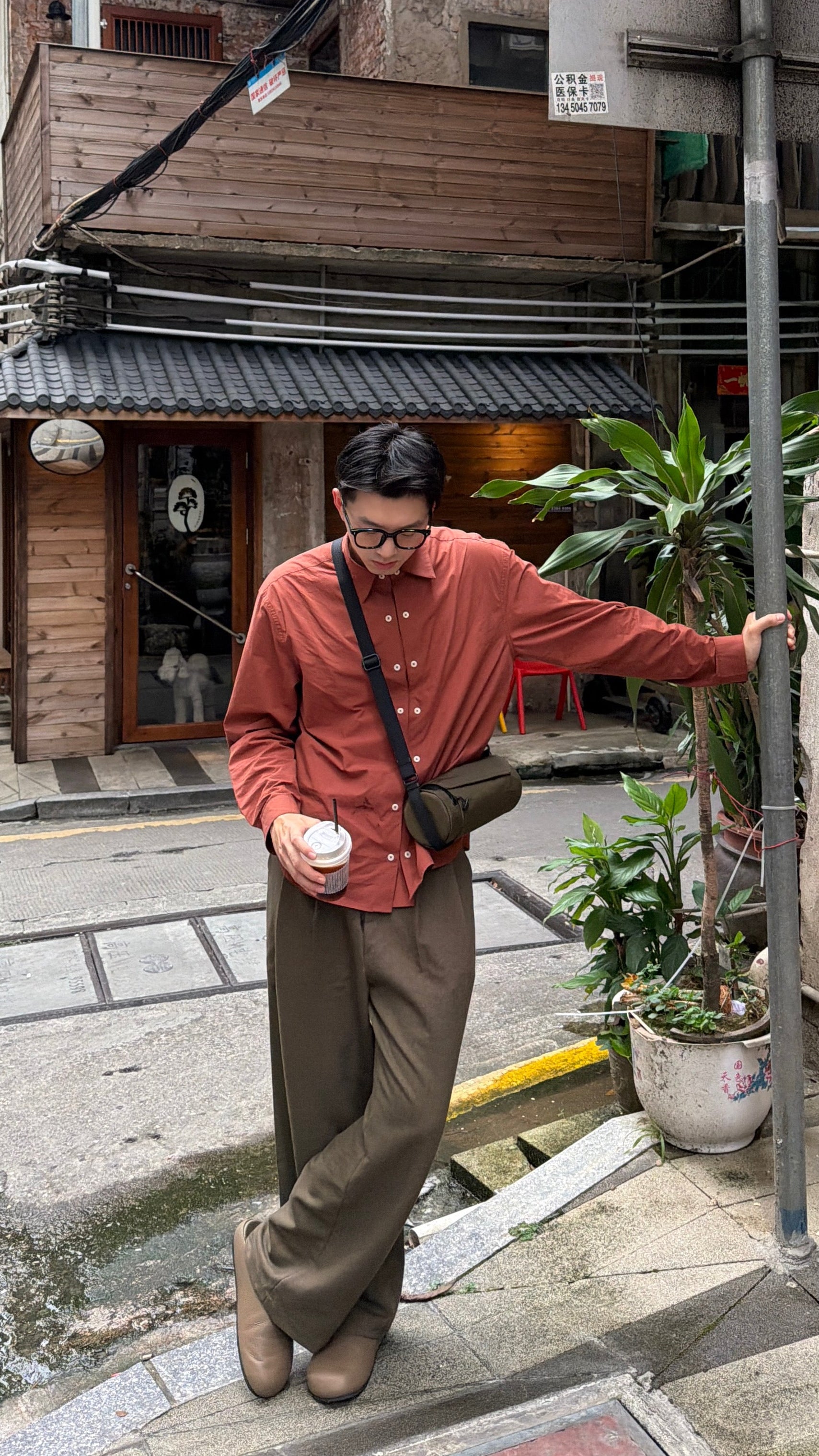 Lifestyle shot: young man holding iced coffee, wearing olive Nayo Smart UL5 Cylinder Sling Bag, compact barrel‑shaped city crossbody bag on city street