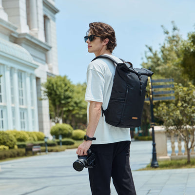 Side view of male model carrying NayoSmart Urban U8 black roll-top backpack with camera on city street, travel commuter lifestyle