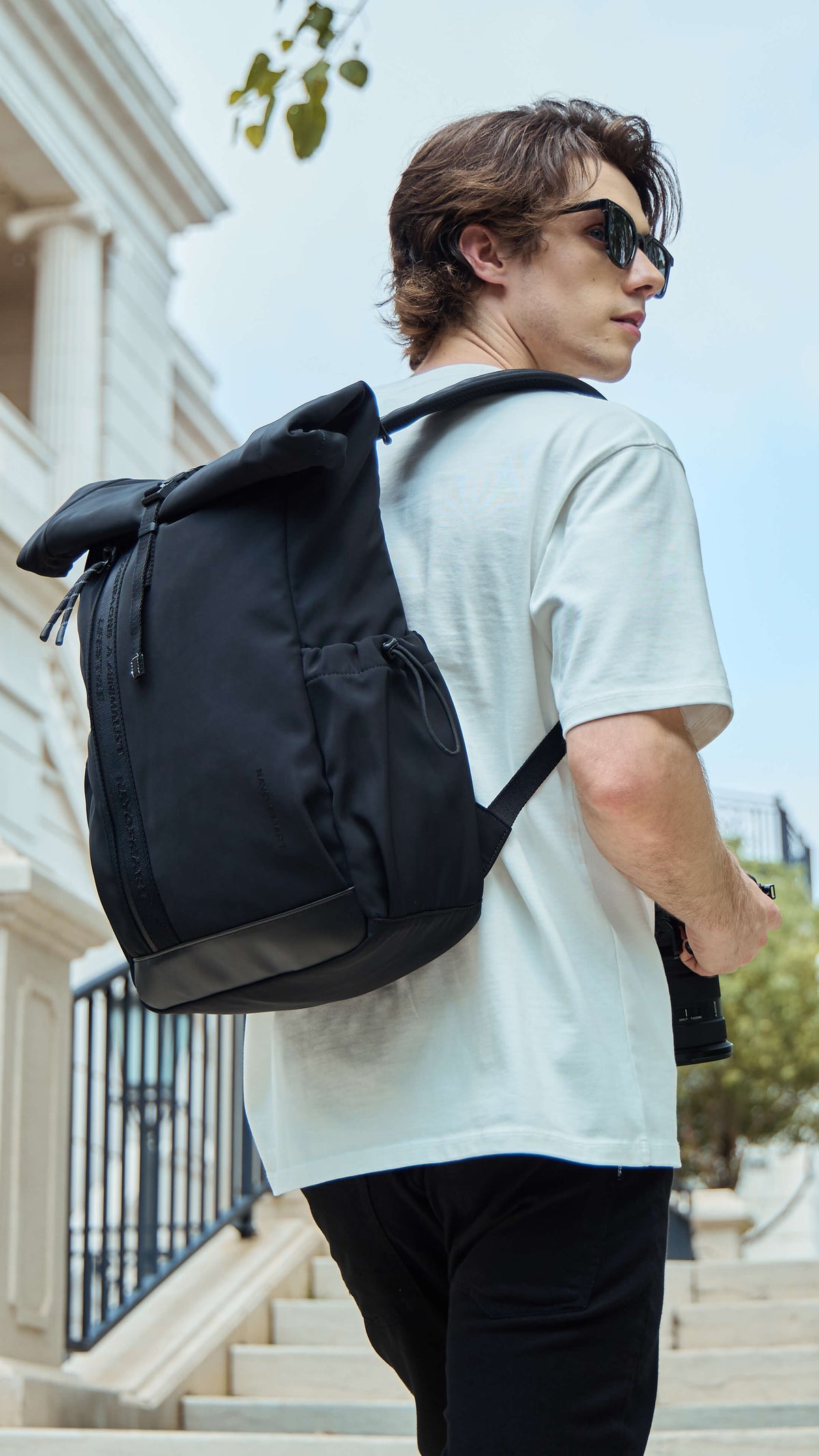 NayoSmart Urban U9 roll-top backpack black model wearing backpack on city street, urban travel photographer holding camera lifestyle shot